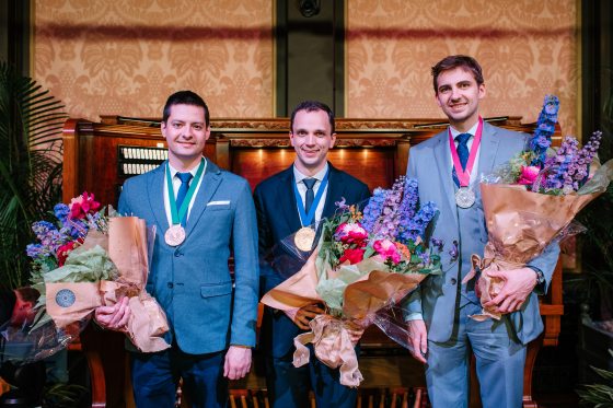 Three winning competitors pose in front of the organ they played, each with a medal around their neck and holding a bouquet of flowers.