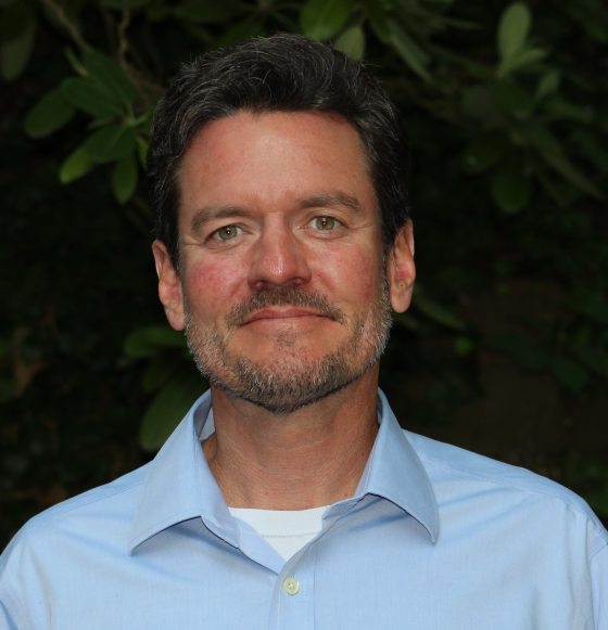 A man in a blue collared shirt poses for a photo against a wall covered in plants