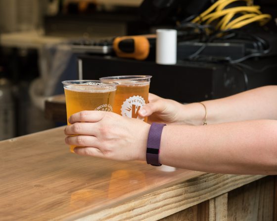 A persons hand taking two plastic cups of beer from a wooden bar. 