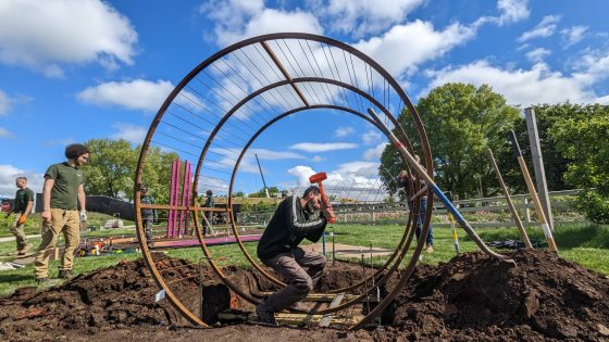 A person using a rubber mallet while installing a circular metal sculpture in an outdoor garden bed.