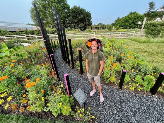 A person in green shirt with straw hat smiling at the camera while standing in a flower garden.