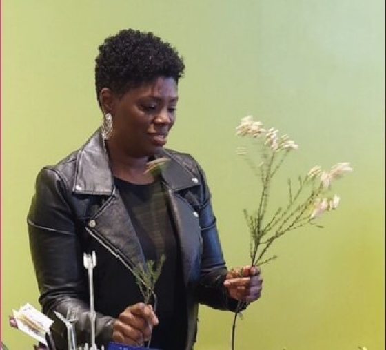 A person with short black hair in a black leather jacket holding a stem of white flowers.