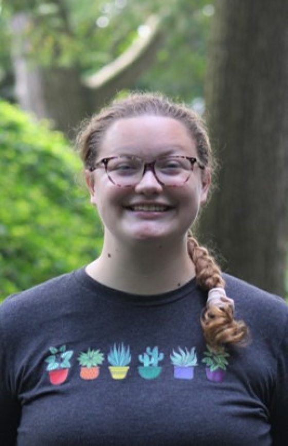 Portrait of a person with light skin and hair pulled back into a long thick braid worn over one shoulder, with an open smile, and wearing eyeglasses and a dark blue tee shirt picturing six small plants in pots, each pot a different color of the rainbow.