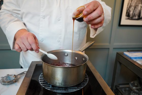 A person in a chef's jacket pouring syrup into a metal pan on a stove.
