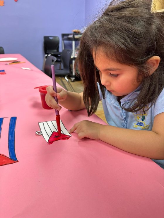 A young child at a table painting a flag with red and black paint.