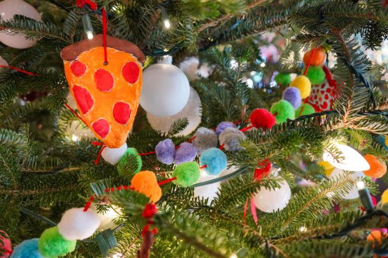A Christmas tree decorated with ornaments made by young children, featuring a pizza slice made of clay and colorful pom-pom garland.