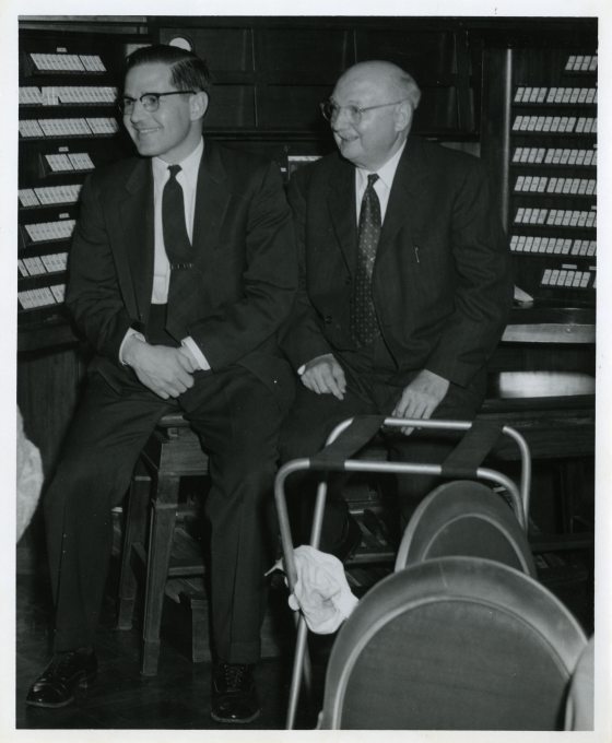 A black and white photo of two people sitting near an organ.