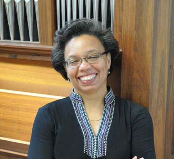 A person with a dark complexion standing in front of an organ, smiling.