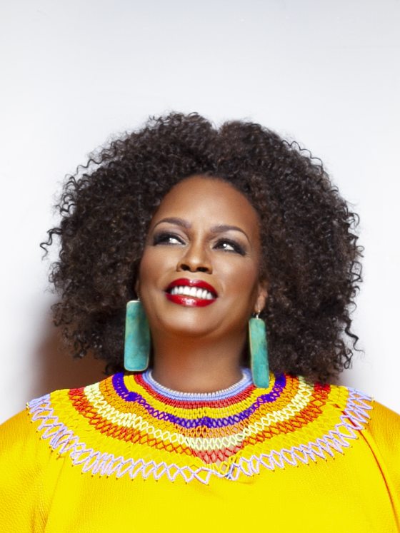 Portrait of a smiling person with bronze skin, black curly hair, red lipstick, dangling green earrings, and a sunshine yellow top, embroidered at the neckline with concentric circles of various colors.