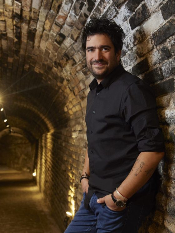 A person with short thick curly hair and beard, wearing a black short-sleeved collared shirt and dark jeans, with an analog watch and tattoos on his left arm, looks at the camera while leaning against the arched brick wall of the interior of a tunnel.