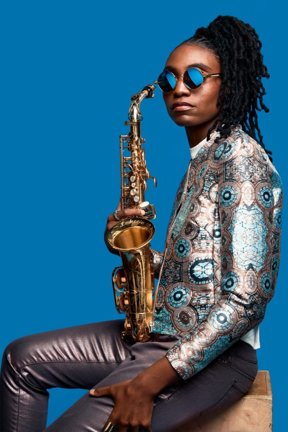 Portrait of a dark-skinned person with black curly hair pulled back, wearing a silver and blue top and silver pants, holding a saxophone, and whose sunglasses seem to reflect the bright blue background.