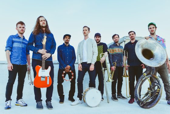 Eight musicians stand and pose for a photo, holding various instruments, including electric guitar, tuba, trombone, trumpet, drum, and tambourine.
