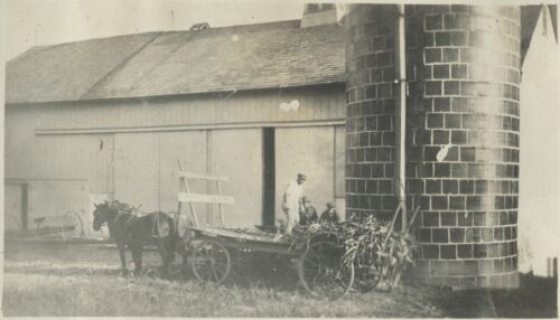 A sepia toned image of a horse pulling a large cart in front of a barn.