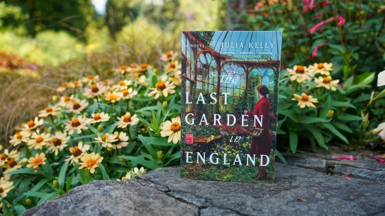 The book "The Last Garden in England" propped up on a stone wall with yellow flowers behind it.