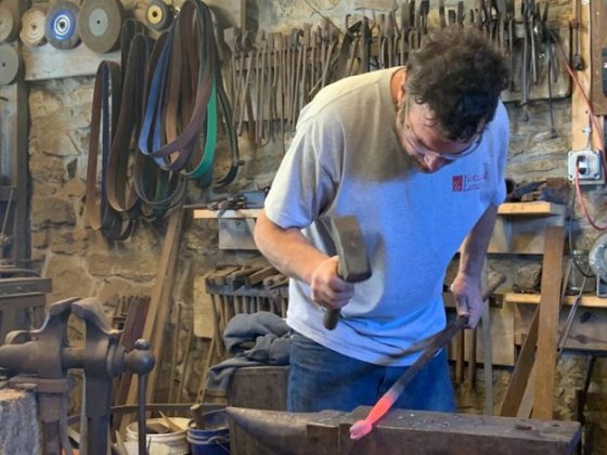 A person using a variety of tools to shape black iron into a snake shape.