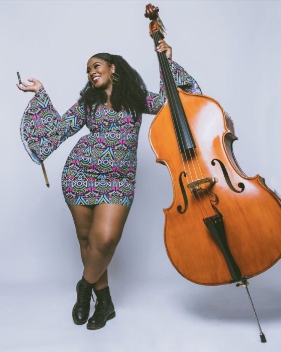 A person with bronze skin, long dark curly hair, broad smile, large earrings, and a colorful geometrically patterned dress strikes a dramatically playful pose holding a bass violin and bow.