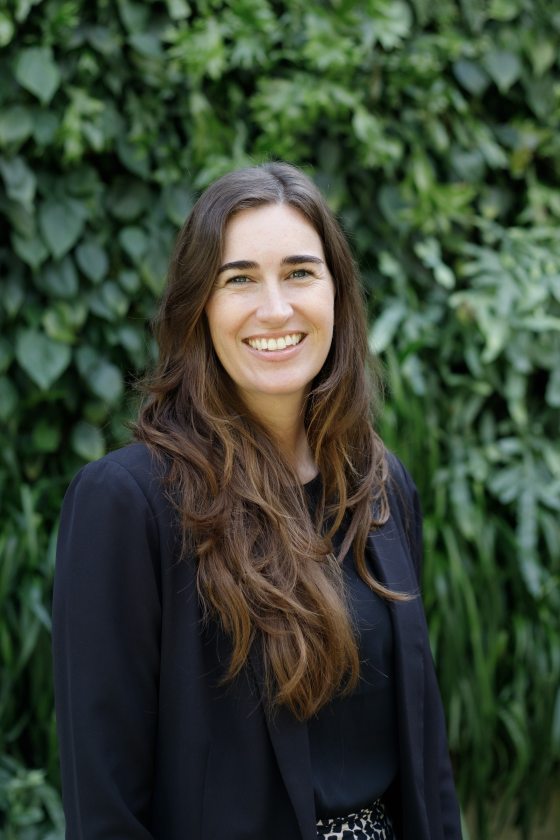 Portrait of smiling person with long brown hair and blue top against a leafy green background.