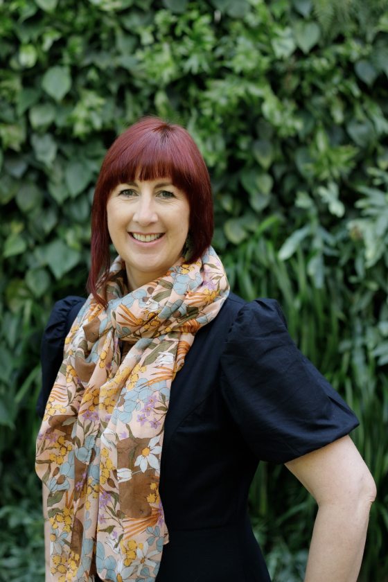 Portrait of person with shoulder-length reddish brown hair and bangs smiling into the camera, wearing a short-sleeve blue top and a long floral neck scarf.