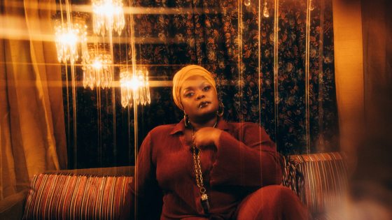 A person wearing a gold headscarf, large hoop earrings, chain necklace, and dark red corduroy outfit looks into the camera while seated on a striped couch with colors in the same palette as their outfit, against a backdrop of dark floral curtains and four small warmly glowing chandeliers.