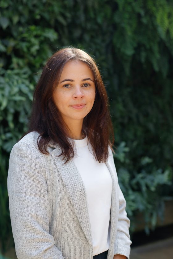 Soft light falls onto the parted brown hair of a person gazing with a pleasant expression toward the camera, wearing a white top and light grayish white herringbone jacket.