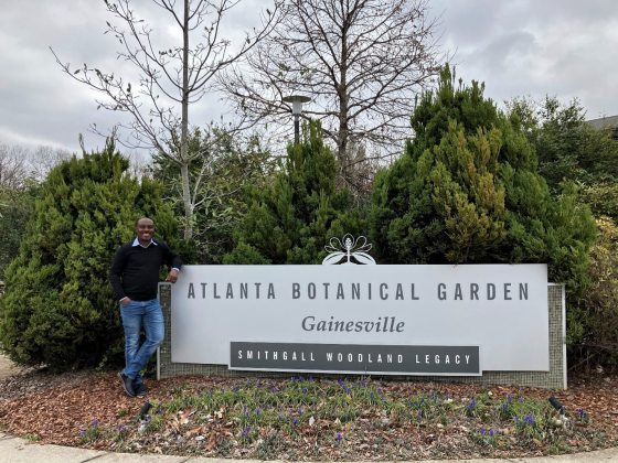 A person with dark complexion standing next to a sign that reads "Atlanta Botanical Garden."
