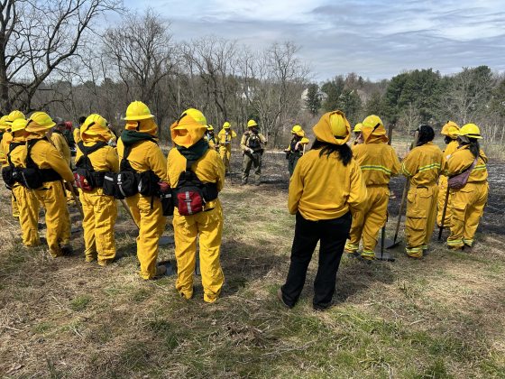 A fire team in yellow protective gear assembled in a circle while in an outdoor field.