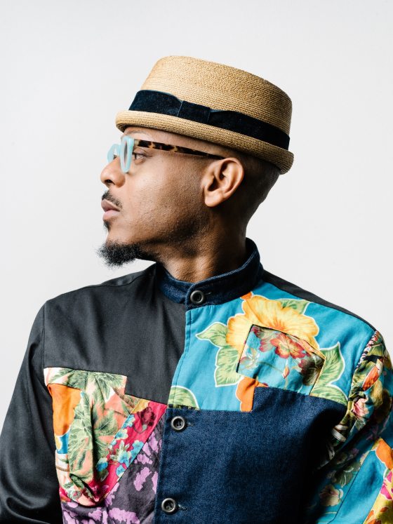 Profile portrait of a person with dark skin, goatee, and mustache, wearing glasses with teal and tortoiseshell frames, a straw hat with black felt band, and a patchwork shirt of shiny black cloth, denim, and colorful floral patches.