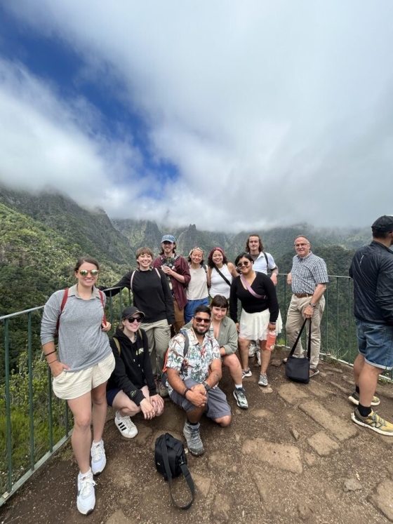 A group of 11 people standing against a railing with mountains behind them smiling at the camera.