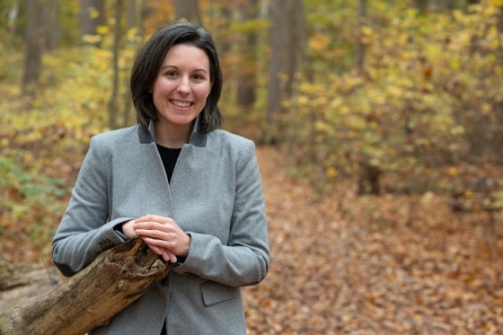 A woman with black hair in a grey jacket stands in a forest and smiles for a photo