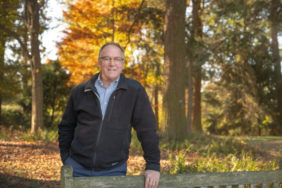 A man with glasses in a zip up sweater poses amongst bright fall foliage 