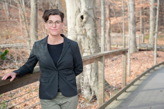 A woman with black hair and glasses with a black blazer rests her arm on a railing on a boardwalk in a forest. 