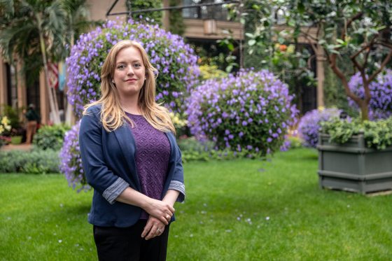 A woman with blonde hair poses for a portrait amid hanging baskets of purple plants.