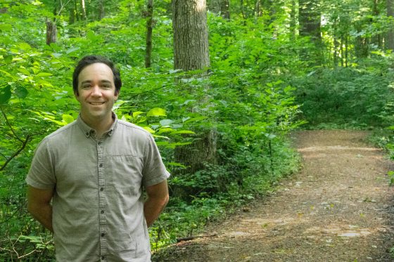 A tan man with thick black hair and a gray button down shirt smiles for a photo in a woodland setting.