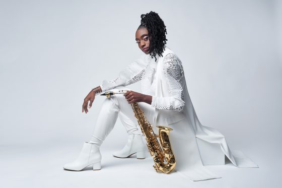 A person with dark complexion in all white sitting on a box with a saxaphone. 