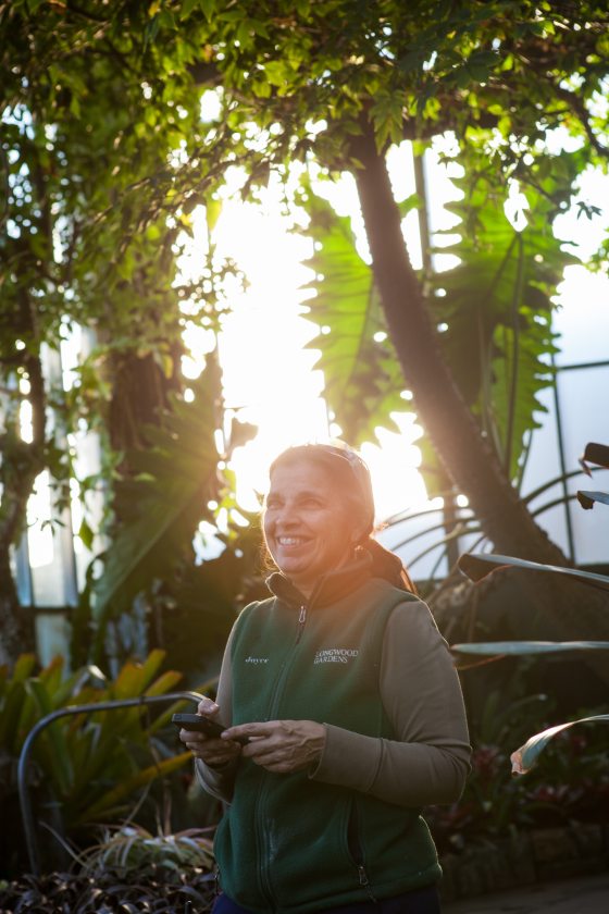 A person smiling and standing in a garden with a halo of sun around them.