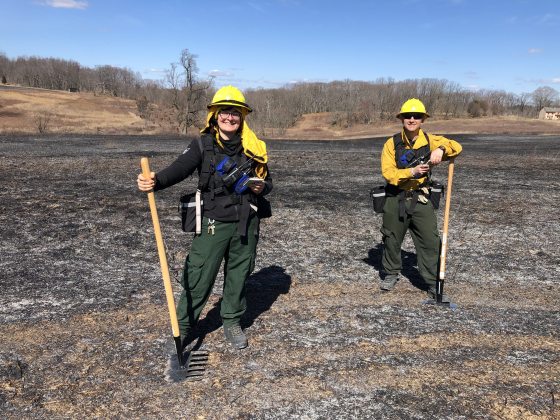 Two people standing in a burned meadow in protective gear.