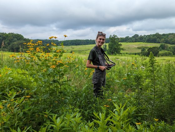 A person standing in tall grass in a meadow smiling at the camera.