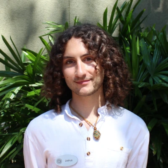 Portrait of person with shoulder-length curly brown hair and light beard, head tilted slightly to viewer's left, wearing white shirt and pendant.