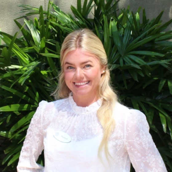 Portrait of person with long blond hair, wearing a lacy white top, smiling at the camera.