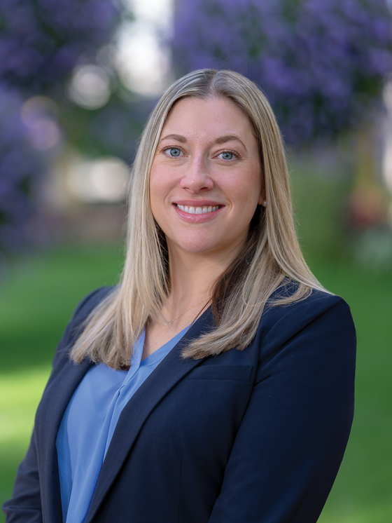 Portrait of smiling person with below-the-shoulder blond hair and blue eyes in a dark blue dress jacket and light blue top against a blurred green and purple garden background.