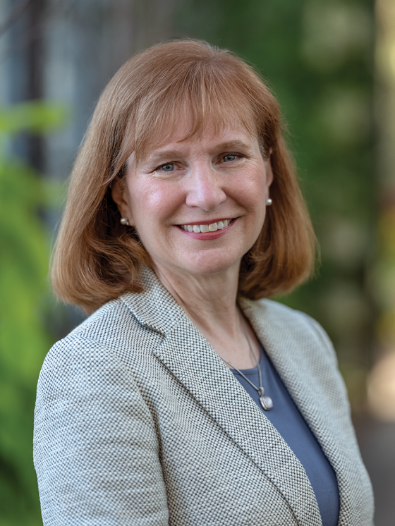 Portrait of person with collar-length light reddish hair and blue eyes smiling at the camera, wearing pearl earrings and pendant, and a gray and white jacket with small hexagonal pattern over a periwinkle-colored tee.