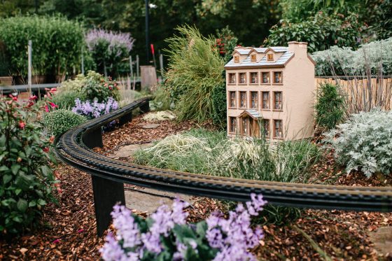 An outdoor train display featuring plantings around the rails and a model of a tan building.
