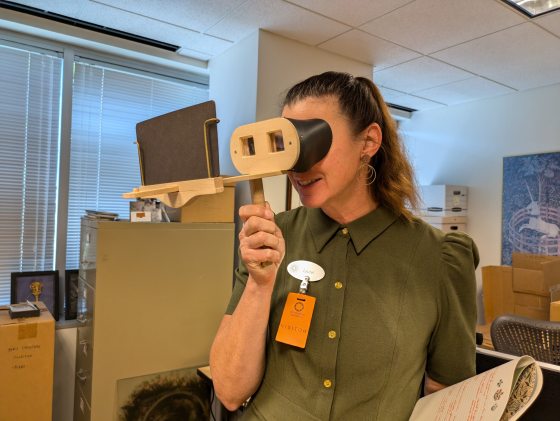 A person in a brown ponytail and hnter green top looking through a stereoscope.