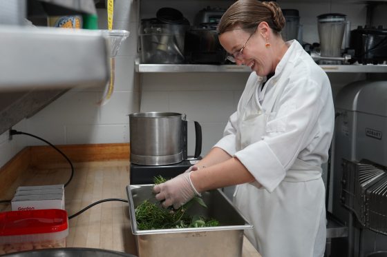 A person in a chef jackets washing herbs in a sink.