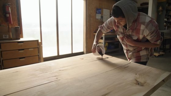 A person in a flannel hoodie pouring a small cup of stain onto a plank of wood.