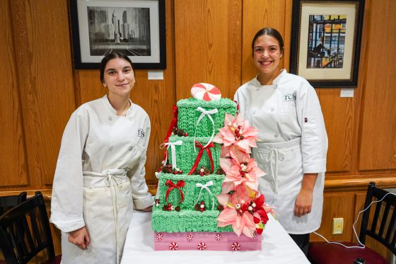 Two people standing beside a holiday cake decorated with green icing and red poinsettia flowers.