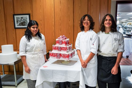 Three people in chef uniforms standing beside a tiered red holiday cake.