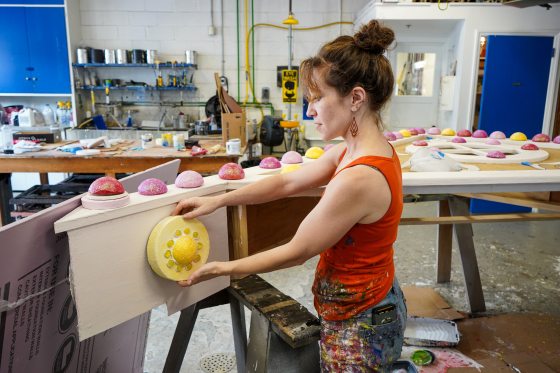 A person holding a yellow, faux, gum-drop, and placing it on the side of a white wood panel.