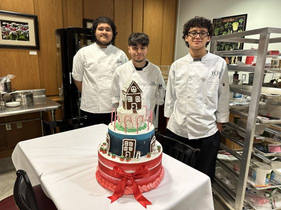 Three people in chef uniforms standing beside a tiered holiday cake., decorated with candy canes, gingerbread, and a red bow.