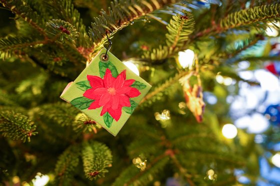 An ornament hanging on a Christmas tree featuring a painted red poinsettia.